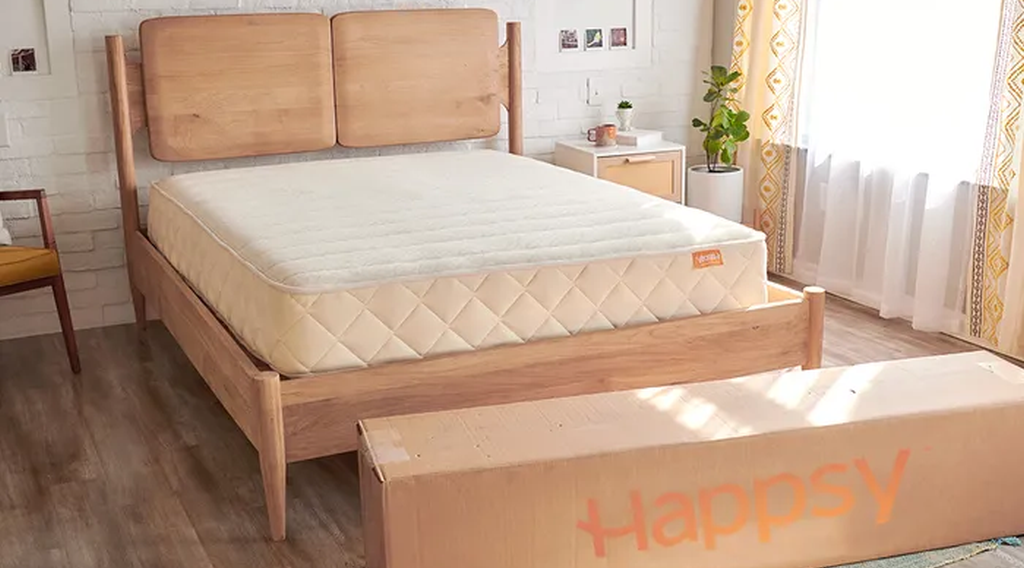 Organic light tan colored Happsy hybrid mattress in a natural wooden frame with shipping box on the floor